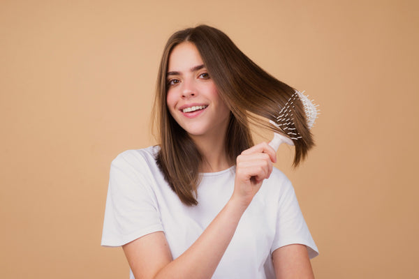Femme brossant ses cheveux bruns mi-longs avec une brosse plate, fond beige — CAMENYS