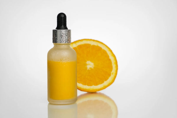 Flacon compte-gouttes de sérum jaune posé à côté d'une tranche d'orange, fond blanc — CAMENYS