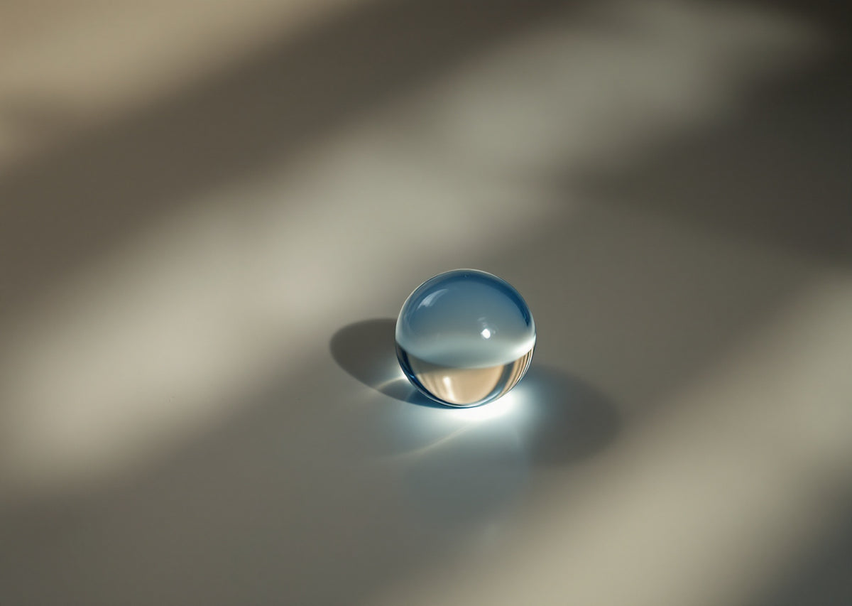 Goutte translucide bleutée posée sur un fond gris pâle rayé par l'ombre d'un store — CAMENYS