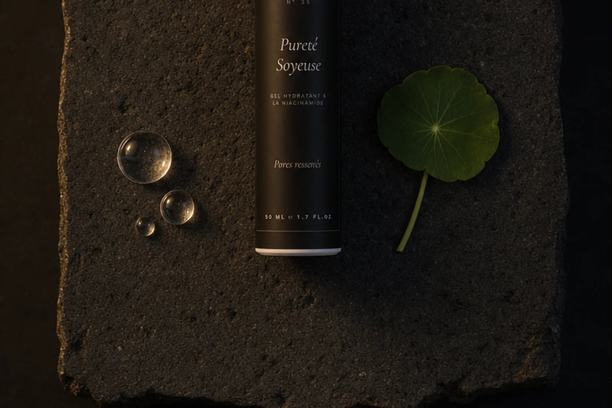 Le flacon noir du gel hydratant à la niacinamide couché sur une dalle de pierre volcanique brune, entre trois gouttes d'eau transparentes et une feuille ronde verte — CAMENYS