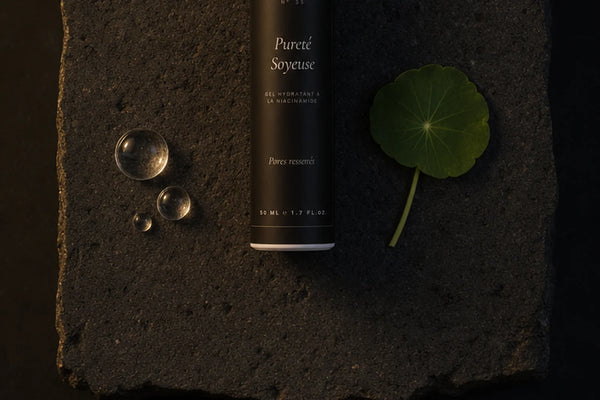 Le flacon noir du gel hydratant à la niacinamide couché sur une dalle de pierre volcanique brune, entre trois gouttes d'eau transparentes et une feuille ronde verte — CAMENYS